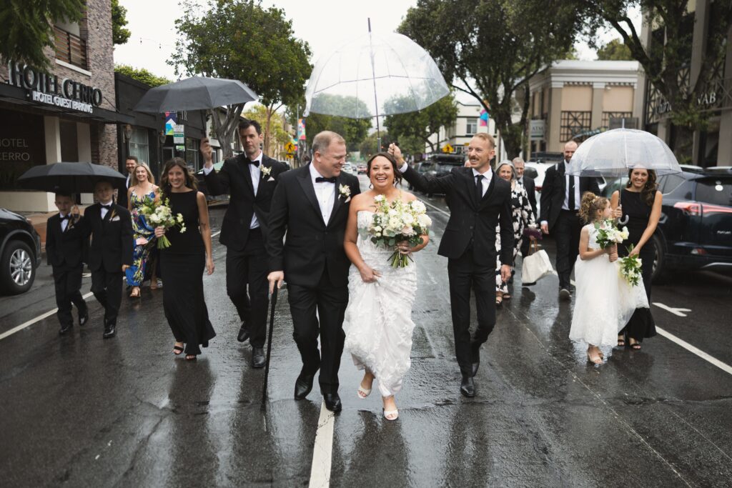 Bride and groom and their wedding party walking down the middle of a rain-soaked street in downtown San Luis Obispo during their wedding day portraits