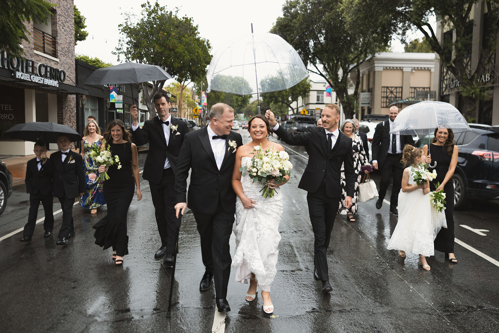 Bride, groom and wedding party walking down a rain-soaked street in downtown San Luis Obispo with clear umbrellas