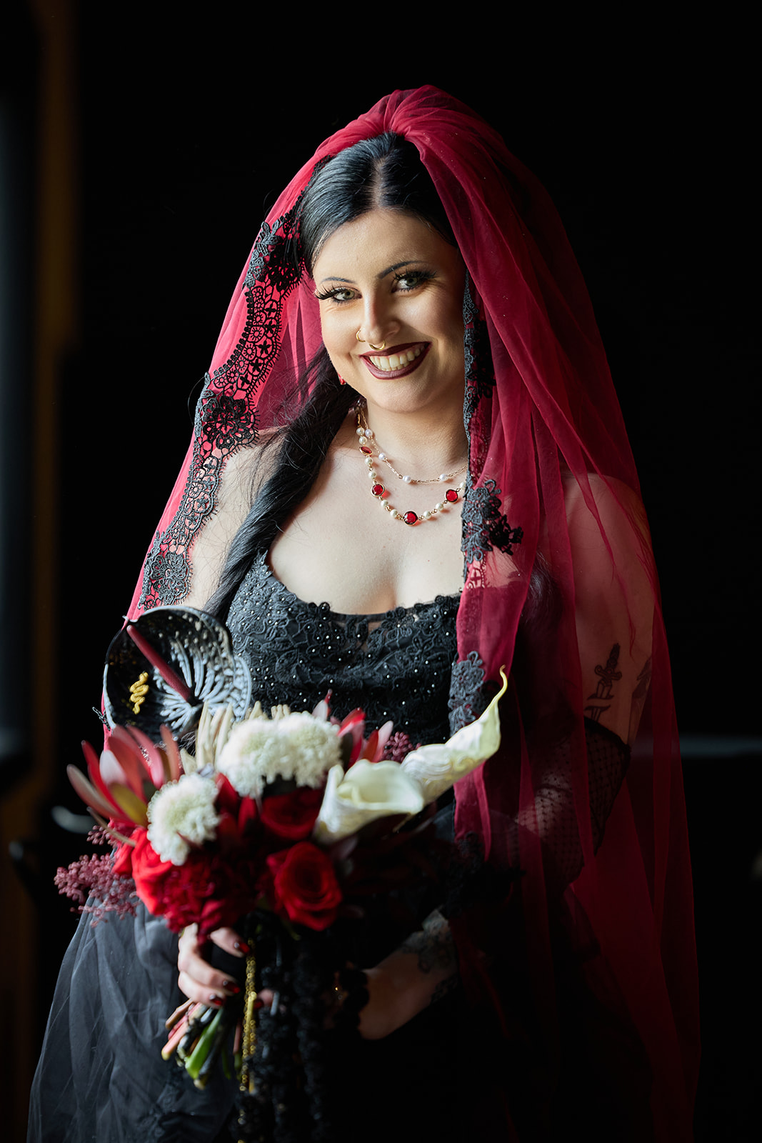 Bride in a black gown and red cathedral veil smiling in dramatic window light, holding red roses and white calla lilies