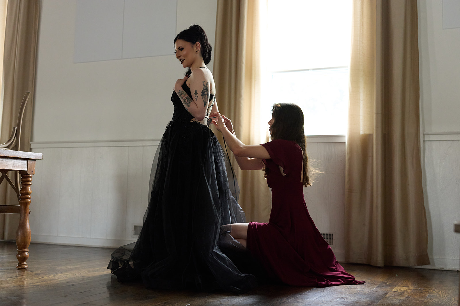 Bride in a black gown at a tall window while her maid of honor adjusts the dress