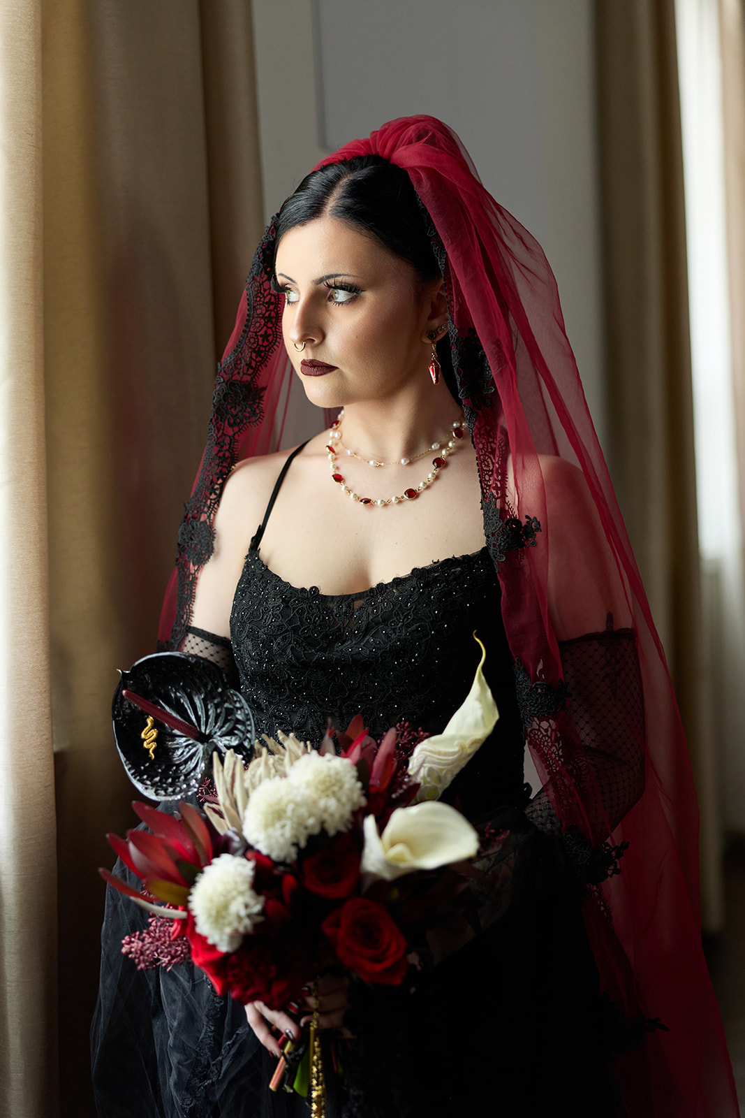 Bride in a red cathedral veil holding a red and white bouquet by the window