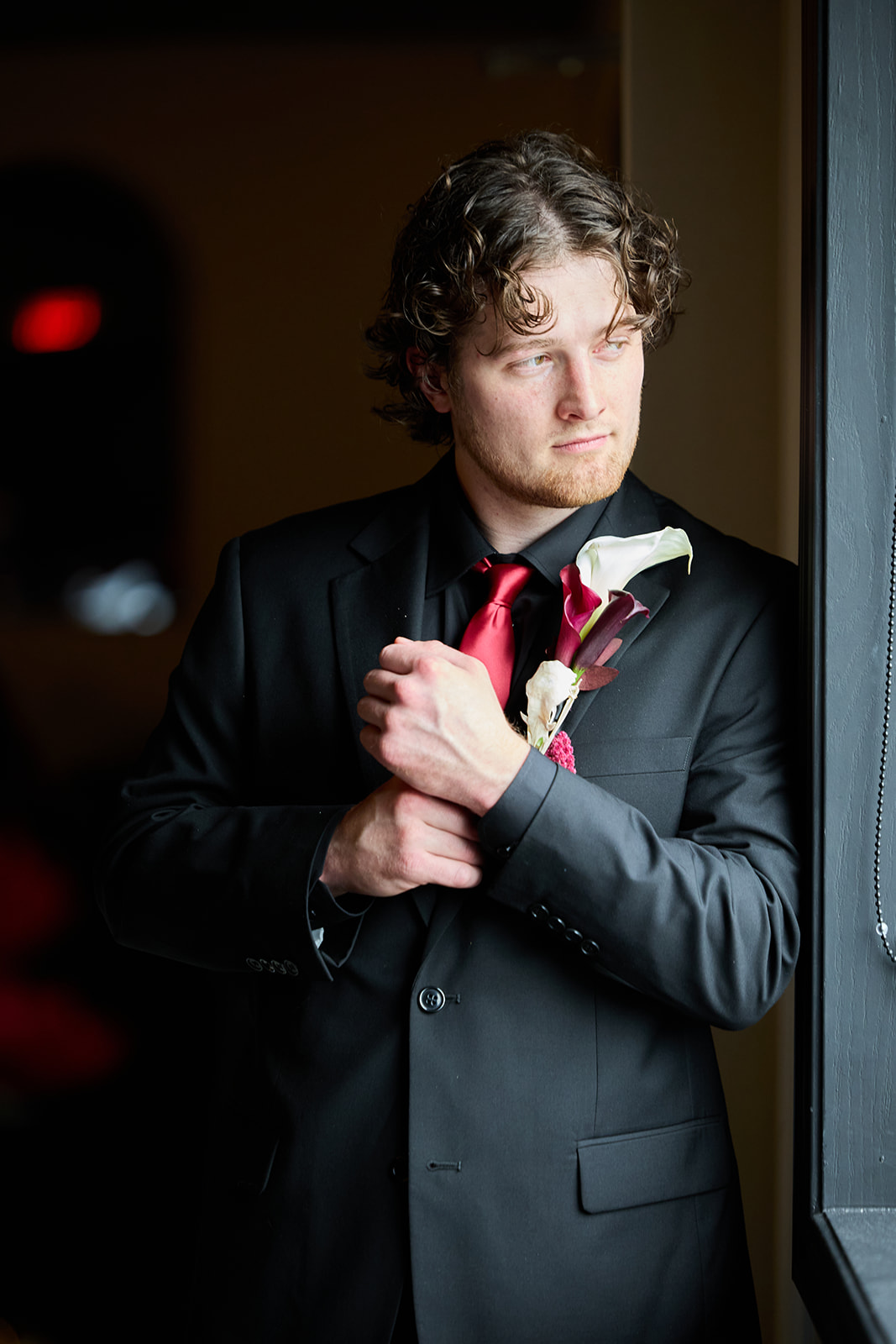 Groom in a black suit and red tie adjusting his cuff by a window