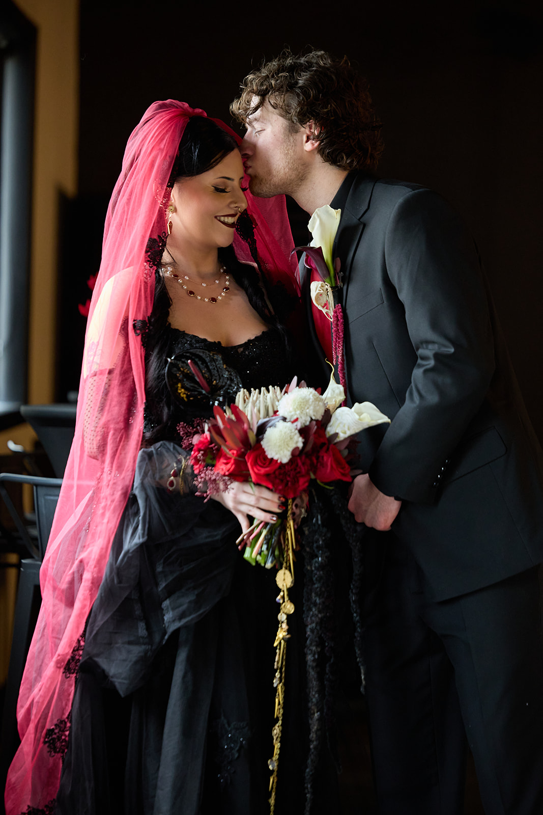 Groom kissing the bride's temple as she holds her bouquet, red veil trailing