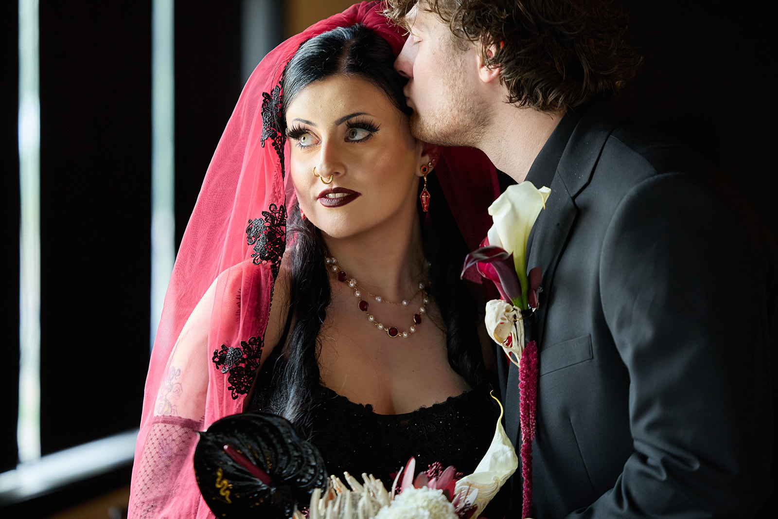 Close portrait of the bride in a red veil with the groom kissing her cheek