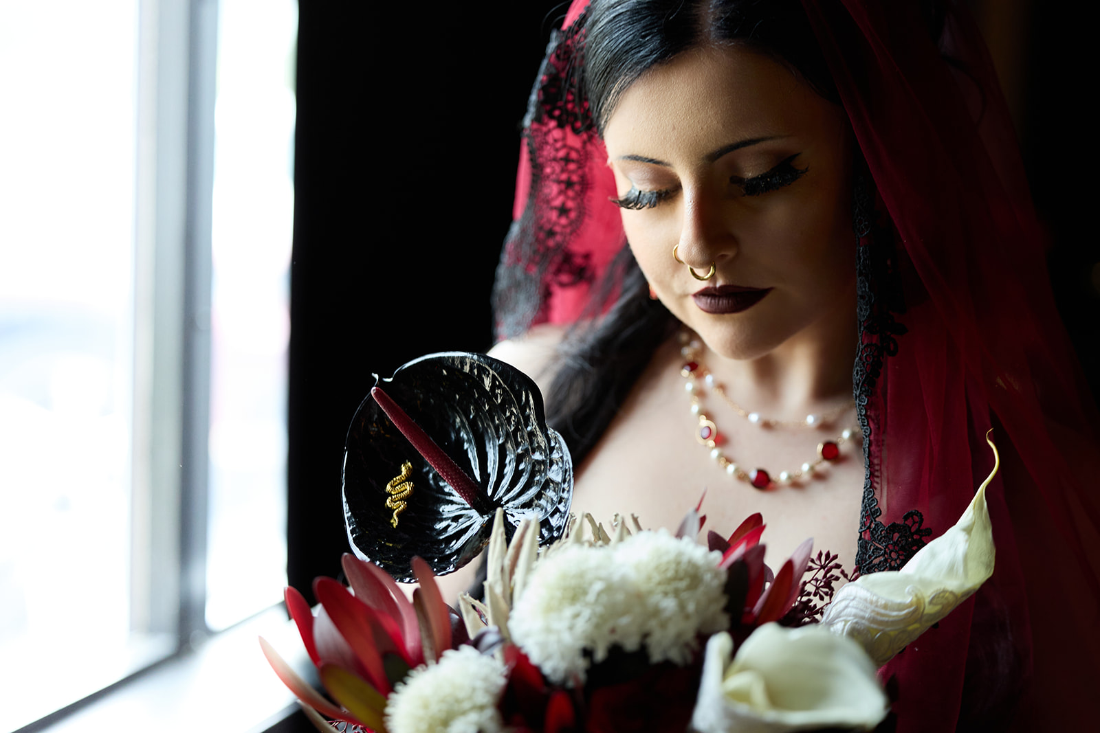 Bride looking down at her bouquet in window light, red veil framing her face