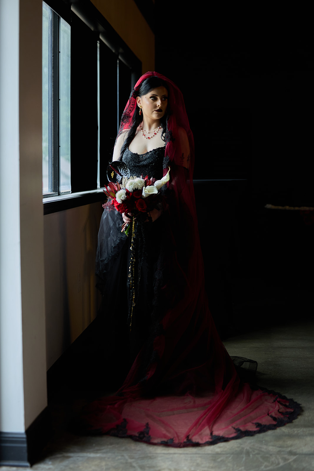 Bride in a black gown with a long red veil standing in a dark hallway by tall windows