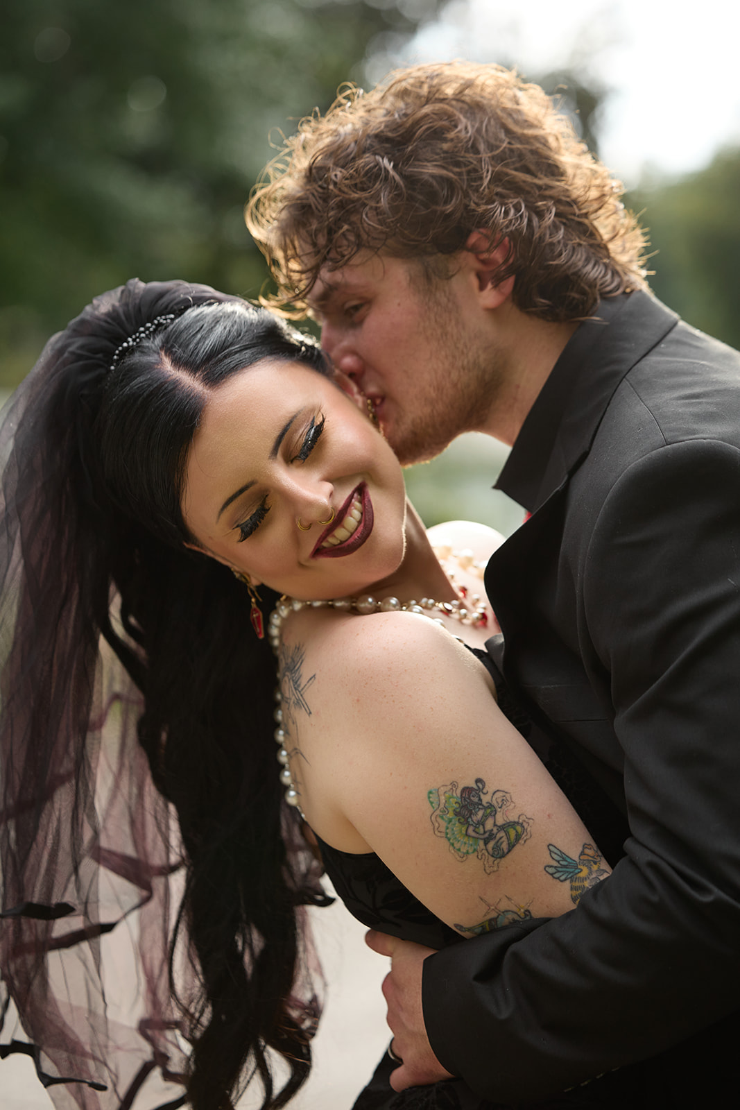 Groom kissing the smiling bride's cheek outdoors, her tattoos showing