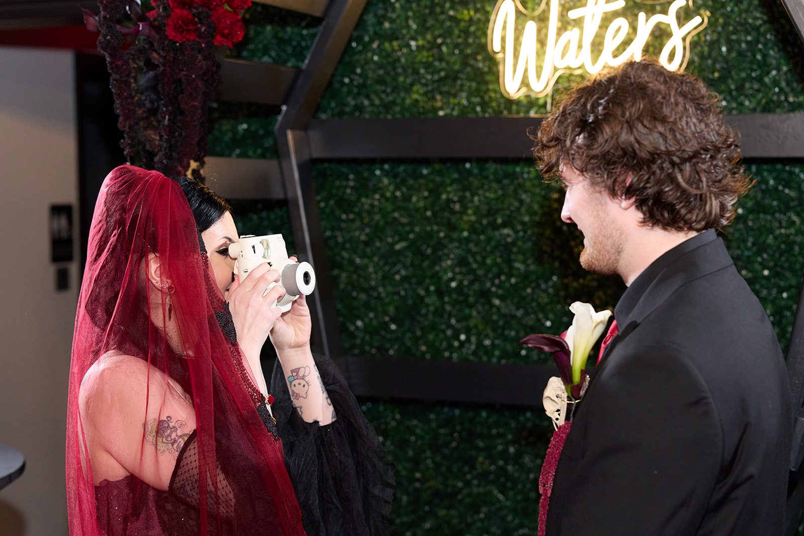 Bride in a red veil taking an instant photo of the groom at the altar under a neon sign