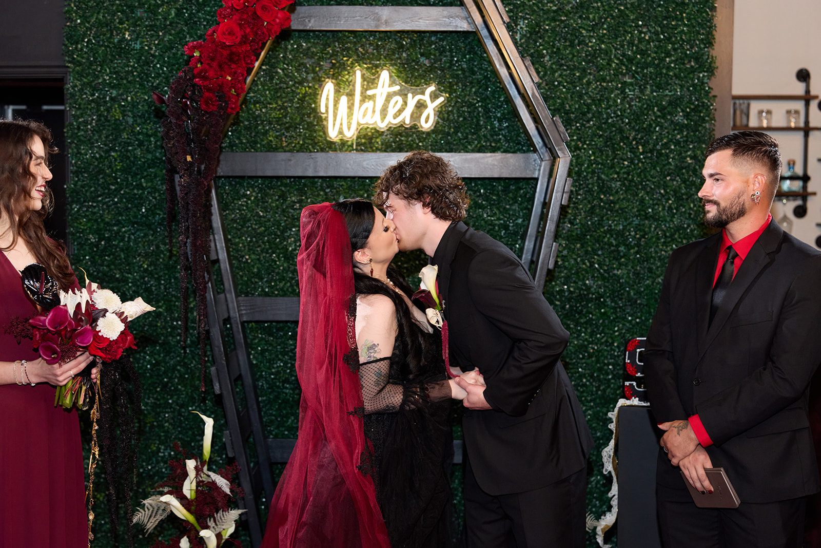 Bride and groom's first kiss in front of a greenery wall with a neon sign